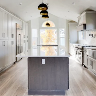 modern kitchen with white cabinetry, a dark island in the foreground, stainless appliances, recessed ceiling lights, and a pendant lamp over the center, with large windows showing autumn trees.